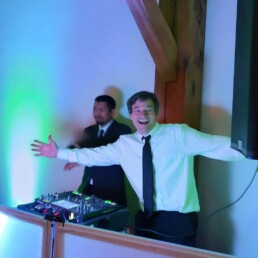 Two professional DJs in suits stand beside their equipment, ready to bring amazing energy to your Vermont wedding. One, a renowned local DJ, beams joyfully with arms open wide, inviting everyone to the dance floor, while his colleague stands behind the table. Vibrant green and blue lights create a festive atmosphere perfect for celebrating your big day.