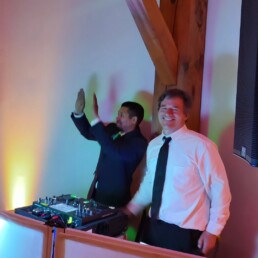 Two men dressed in formal attire stand beside professional DJ equipment; one smiles warmly at the camera, while the other, likely a sought-after Vermont wedding DJ, is caught mid-celebration—clapping or dancing. Vibrant lights illuminate the background, setting a festive mood perfect for your special day.