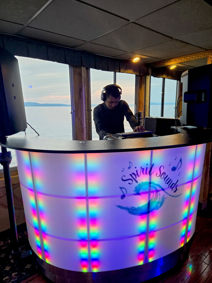 A DJ from Spirit Sounds mixes music behind a glowing booth on a boat, with sunset views of Vermont mountains and water—ideal for weddings.