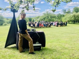 Vermont DJ Emma Manion stands by her professional DJ setup on a lush grassy field, beaming warmly at the camera. Behind her, guests are seated outdoors, watching a beautiful wedding ceremony unfold beneath a rustic wooden arch on a radiant sunny day—the perfect setting for your unforgettable Vermont celebration.