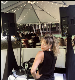 Vermont wedding DJ Emma Manion stands at her DJ booth inside a beautifully lit tent, warmly smiling at the camera. Behind her, guests enjoy their evening seated at elegantly arranged tables beneath romantic string lights, while professional speakers on either side promise an unforgettable celebration.