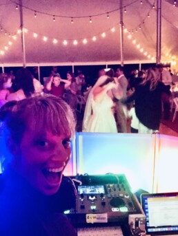 A joyful Vermont DJ captures a selfie at a lively wedding reception, surrounded by vibrant lights and professional DJ equipment, as guests and the bride dance beneath romantic string lights—setting the perfect atmosphere for an unforgettable celebration.