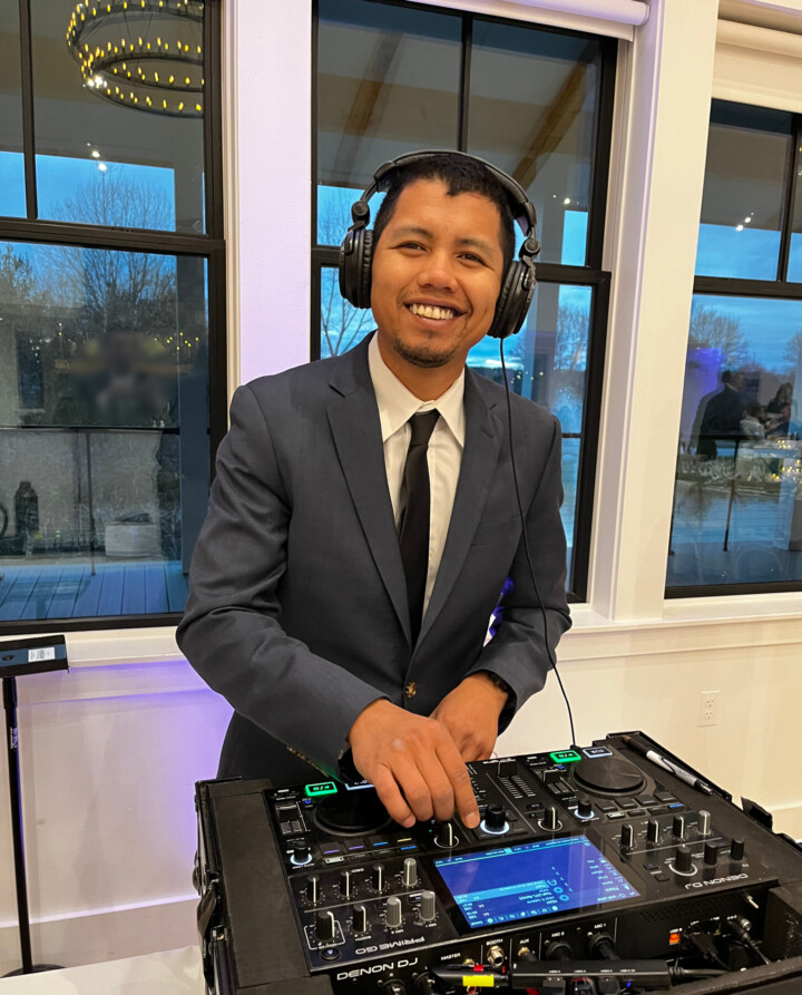 A smiling, well-dressed DJ mixes music indoors as soft evening light and Vermont trees shine through large windows—perfect wedding vibes.