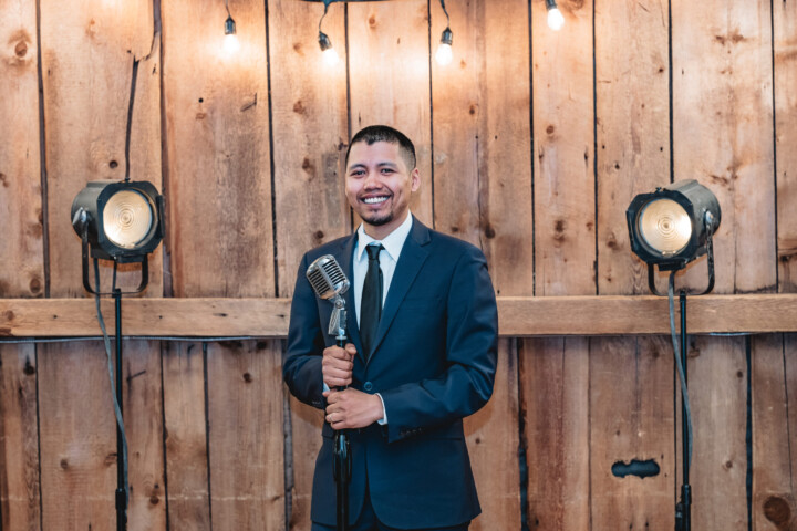 A smiling DJ in a sharp suit holds a vintage mic under string lights, ready to bring charm and fun to your Vermont wedding.