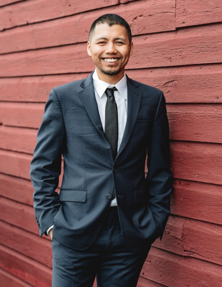 A friendly Vermont wedding DJ in a dark suit and tie smiles confidently by a red wooden wall, ready to make your day unforgettable.