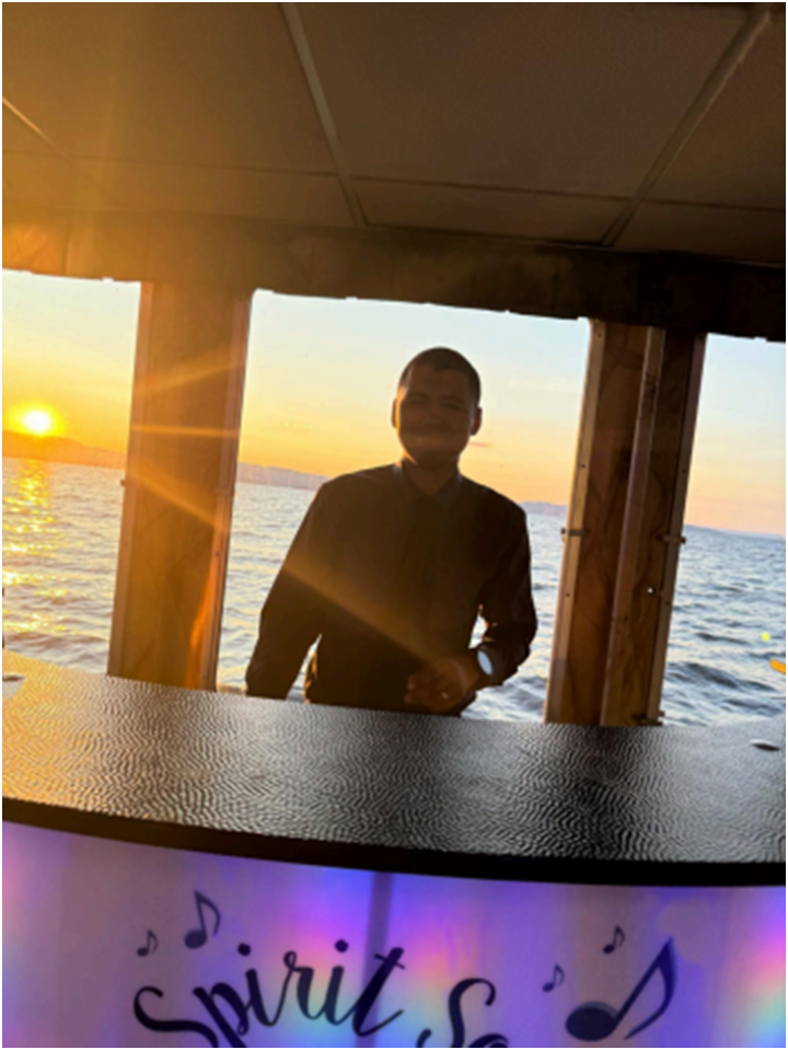 A DJ stands behind a vibrant, Vermont-style wedding setup on a boat at sunset, colorful lights glowing and 