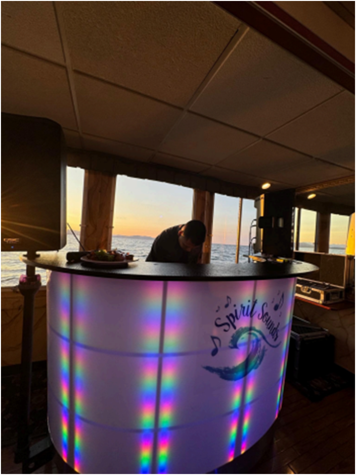 A DJ stands behind a glowing Spirit Sounds bar on a sunset boat, calm Vermont waters outside—perfect for your wedding celebration.