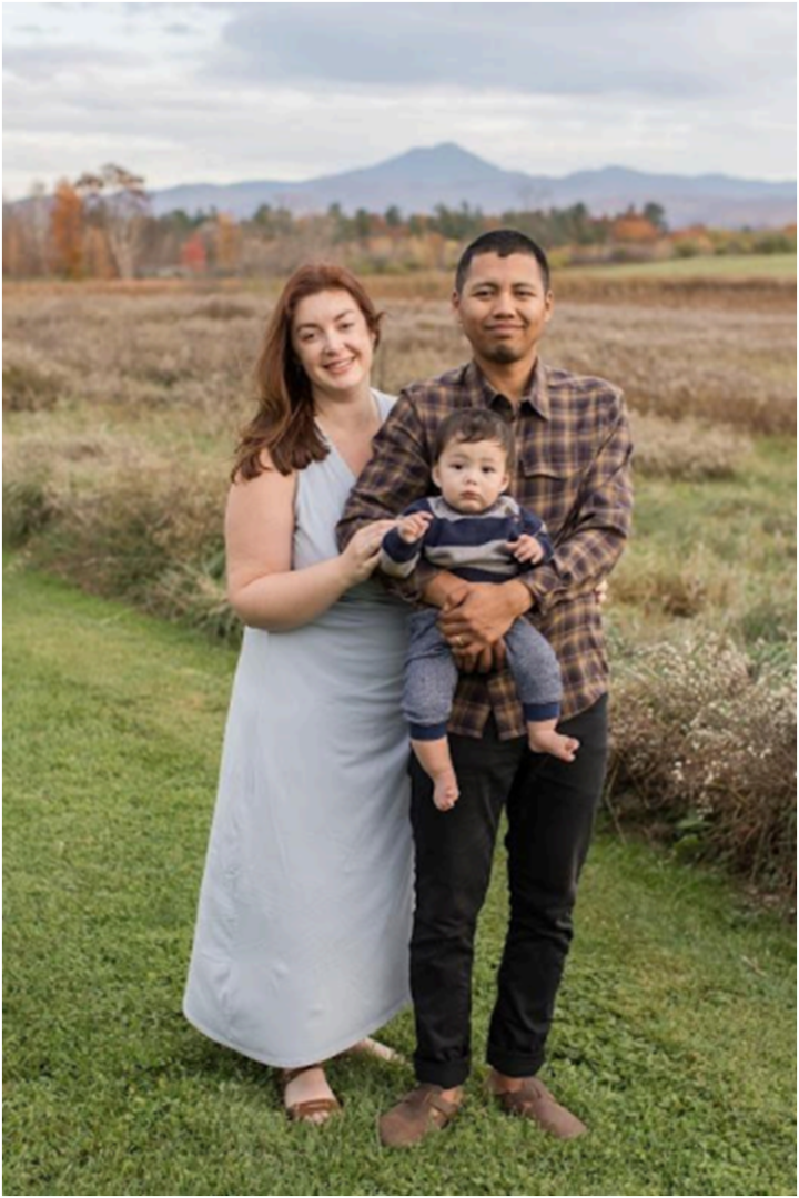 A happy couple and their baby pose on grassy fields with Vermont’s mountains—ready for unforgettable moments with a local wedding DJ.