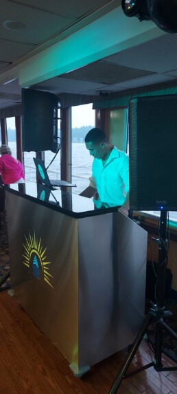 Vermont wedding DJ Aaron Carroll, dressed in a crisp white shirt, stands behind an elegant booth featuring a sun logo as he mixes music on his laptop. Large speakers promise crystal-clear sound while bright windows overlook a scenic water view—creating the perfect romantic atmosphere for your special day. Another guest can be seen enjoying the background ambiance.