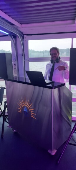 Vermont wedding DJ Dustin Bruley, dressed in a crisp white shirt and tie with headphones on, stands behind his signature sunburst-logo DJ booth, using his laptop to set the perfect mood. Large speakers ensure crystal-clear music, while a picturesque window view of sparkling water and lush trees creates an inviting celebration backdrop.