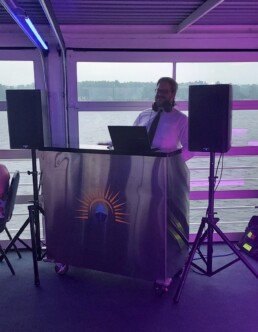 A professional DJ, dressed in a tie and headphones, stands behind a sleek metallic booth flanked by speakers. Through large windows, couples can admire serene water and trees bathed in the romantic purple glow of dusk—a magical atmosphere often created by Vermont wedding DJ Dustin Bruley.