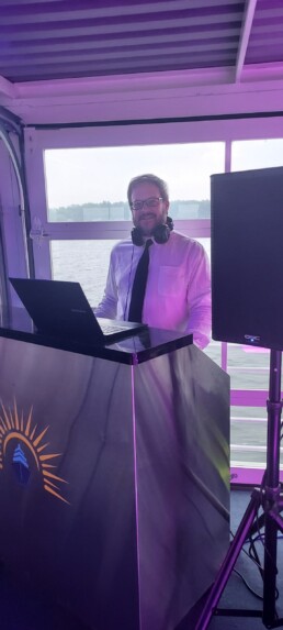 Vermont wedding DJ Dustin Bruley, dressed in a crisp white shirt and headphones, stands behind a DJ booth with a laptop, smiling warmly. Ready to create an unforgettable celebration for your special day, Dustin is set up with professional equipment and picturesque water views visible through the window behind him.