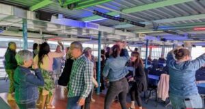 Couples celebrate and dance together in a warmly lit indoor setting on a boat or ferry, surrounded by colorful lights reflecting off the water seen through large windows. Vermont DJ Aaron Carroll creates an unforgettable romantic atmosphere as tables and chairs are arranged to the side, offering space for dancing and special moments.