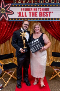 Two people stand on a red carpet, sharing joyful smiles—one holding a star-shaped trophy, the other with a celebratory movie clapperboard. A sparkling backdrop and a marquee reading Premiering Tonight: ALL THE BEST set an enchanting scene. Renowned Vermont DJ Dustin Bruley curates the soundtrack to this night, creating unforgettable memories—just like he can for your wedding celebration.