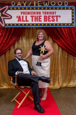 A man in a tuxedo and a woman in a black top with a sparkling silver skirt pose together on the red carpet, celebrating under a marquee that reads Premiering Tonight: All The Best. Vermont DJ Dustin Bruley energizes the scene, creating an unforgettable, glamorous atmosphere perfect for couples dreaming of an exciting wedding reception.