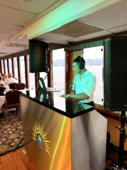 Vermont wedding DJ Aaron Carroll, wearing headphones, stands behind a stylish booth adorned with a sun logo, expertly mixing on his laptop and DJ controller aboard a boat with expansive windows offering stunning river views—creating the perfect ambiance for your special day.