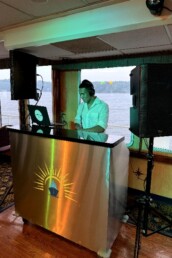 Vermont DJ Aaron Carroll, wearing headphones, expertly mixes music behind a sleek DJ booth adorned with a sun logo, creating an unforgettable atmosphere on a boat with large windows showcasing scenic water and lush trees&mdash;an ideal backdrop for couples dreaming of a unique and memorable wedding celebration.