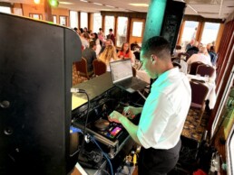 A Vermont wedding DJ in a crisp white shirt expertly mixes music on a controller during a joyful indoor celebration, with happy guests seated at elegantly decorated round tables in the background.