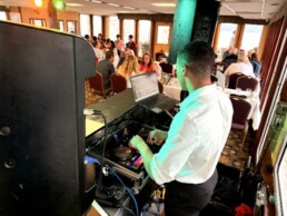 A Vermont wedding DJ in a crisp white shirt expertly mixes music beside a laptop at an elegant indoor reception, setting the perfect mood as guests enjoy themselves at beautifully arranged round tables in the background.