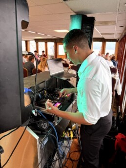 A Vermont wedding DJ in a crisp white shirt expertly manages a mixing console at an elegant indoor venue, with happy guests seated at beautifully arranged tables in the background.