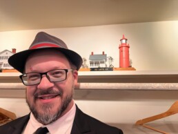 A friendly man in a suit and fedora flashes a welcoming smile in front of a display shelf featuring a quaint model house and a red lighthouse labeled “Isle La Motte 1827,” exuding the inviting, vintage-inspired charm Vermont couples love for their unforgettable wedding celebrations.
