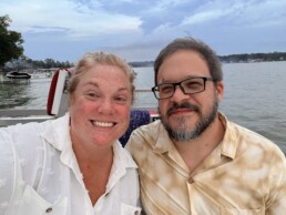 A joyful couple sits together on a boat, surrounded by a peaceful lake and lush trees under a partly cloudy sky, soaking in the romantic atmosphere after choosing Dustin Bruley as their Vermont wedding DJ.
