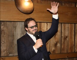 Vermont wedding DJ Dustin Bruley, dressed in a sharp suit and glasses, holds a microphone and raises his hand while speaking warmly to the crowd. Standing before a rustic wooden wall under soft round lighting, he exudes charisma and professionalism—ready to create an unforgettable celebration for couples on their special day.