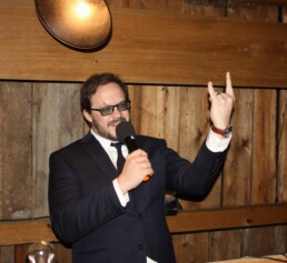 Vermont wedding DJ Dustin Bruley, dressed in a sharp suit and glasses, stands enthusiastically in front of a rustic wooden wall. He holds a microphone and raises his hand in a fun 'rock on' gesture, ready to bring energy and excitement to your special day.