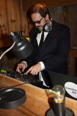 A professional Vermont DJ in a suit and headphones—possibly Dustin Bruley—expertly mixes music on his console inside a warm, wooden venue. A softly glowing lamp, laptop with SuperSound DJ sticker, and additional equipment sit neatly on the desk, creating an inviting atmosphere perfect for weddings.
