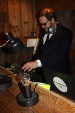 A Vermont wedding DJ, dressed in a suit and headphones, expertly mixes music from a stylish wooden booth. A glowing lamp and modern lightbulb add romantic ambiance to the counter in the foreground, creating the perfect setting for your special day.