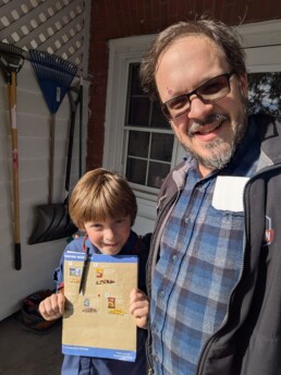 A smiling child in a scouting uniform holds up a menu sheet next to an adult, both standing outdoors by a brick wall with garden tools hanging behind them. Sunlight casts gentle shadows on their faces as they share a joyful moment together. This heartwarming scene reflects the spirit of connection and celebration that Vermont DJ Dustin Bruley brings to every wedding, creating unforgettable memories filled with music and warmth for couples on their special day.
