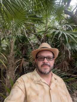 Vermont DJ Dustin Bruley, wearing glasses, a straw hat, and a tan shirt, smiles for a relaxed selfie in front of lush green tropical foliage—a welcoming and personable DJ ready to bring fun and great music to your special day.