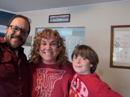 Three people, two adults and a child, smiling indoors and dressed in matching red shirts, stand close together. Behind them are a framed document and painting that create a welcoming atmosphere—capturing the kind of joyful family moment you could experience at your own Vermont wedding celebration with DJ Dustin Bruley providing the perfect soundtrack.