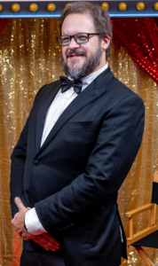 A smiling man with glasses and a beard, dressed in a sharp black tuxedo and bow tie, stands against a sparkling gold and red backdrop with his hands clasped—Vermont wedding DJ Dustin Bruley, ready to create an unforgettable celebration for your special day.