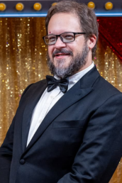 A friendly, bearded man with glasses beams in a sharp tuxedo and bow tie, standing before a sparkling gold glitter curtain accented with red and blue decorations&mdash;highlighting the welcoming and professional presence Vermont DJ Dustin Bruley brings to wedding celebrations.