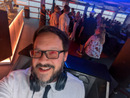 Vermont DJ Dustin Bruley, wearing glasses and headphones around his neck, takes a cheerful selfie on a boat or ship where guests mingle near a bar with stunning water views through large windows—a scene radiating the relaxed, joyful atmosphere perfect for unforgettable wedding celebrations.