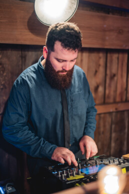 A bearded DJ in a blue shirt stands indoors, expertly managing a DJ controller amid warm lighting and rustic wooden walls—the perfect atmosphere for creating unforgettable wedding moments in Vermont.