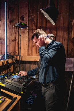 A professional DJ in a blue shirt expertly mixes music in a cozy, wooden venue filled with warm lighting—perfect for an unforgettable Vermont wedding celebration—while surrounded by quality audio gear and thoughtfully adjusting controls with headphones.