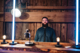 A bearded man in a blue shirt and black tie stands ready to entertain behind a rustic wooden DJ booth, illuminated by soft blue vertical lights and warm exposed bulbs, creating a romantic atmosphere inside a cozy wood-paneled venue—perfect for Vermont couples seeking an unforgettable wedding celebration.