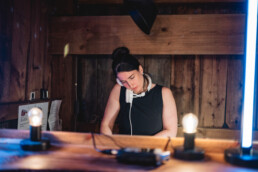 A stylish woman in a black dress stands at a wooden counter, confidently using a vintage rotary phone, headphones resting around her neck to highlight her DJ expertise. On the counter in front of her are glowing light bulbs and professional electronic equipment—setting the stage for an unforgettable wedding celebration in Vermont.