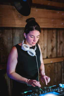 A professional female DJ in a sleeveless black top with white headphones around her neck expertly works her DJ controller indoors, creating the perfect atmosphere under warm lighting. She stands in front of a charming wooden wall, ideal for couples seeking an inviting Vermont wedding vibe.